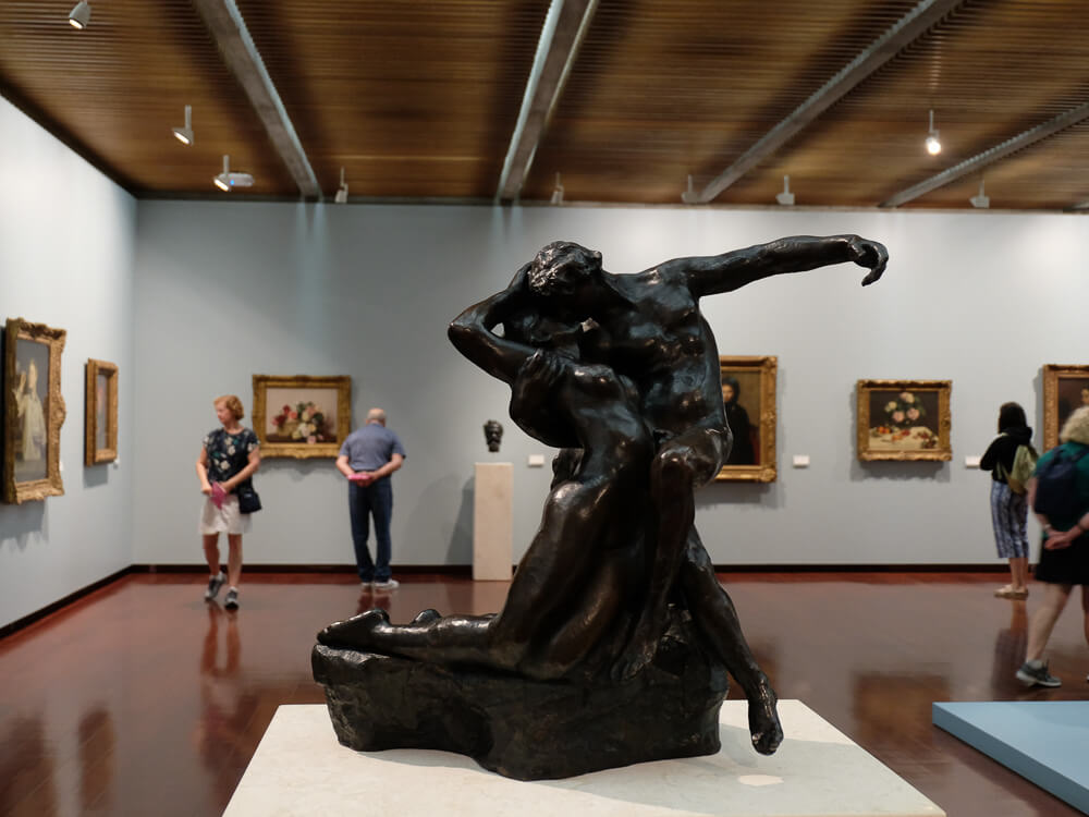 Interior of an art gallery showcasing classical paintings and a prominent bronze sculpture of two intertwined figures.