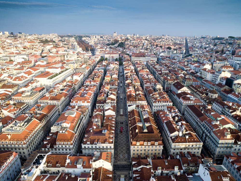 Aerial view of Baixa, the central district of Lisbon, showcasing its distinctive grid layout. The image highlights wide, straight streets flanked by rows of buildings with red-tiled roofs. The district is framed by the cityscape under a clear blue sky.