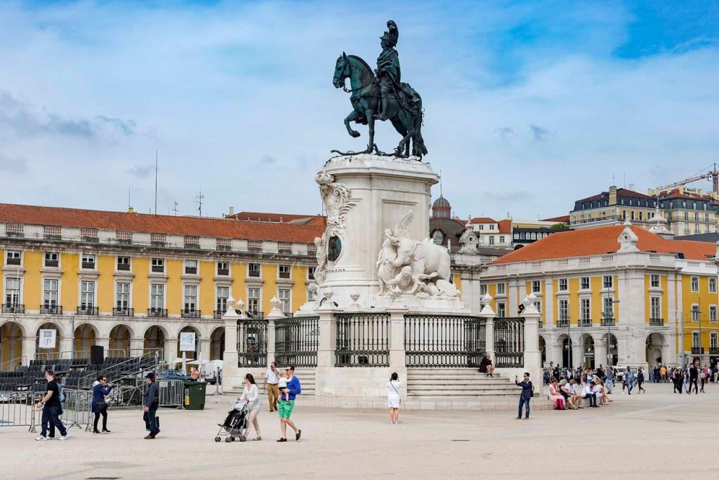 The Praça do Comércio in Lisbon, featuring a grand statue of King José I on a horse at the center. The square is bordered by yellow buildings with arcades, and people are seen walking and enjoying the open space.