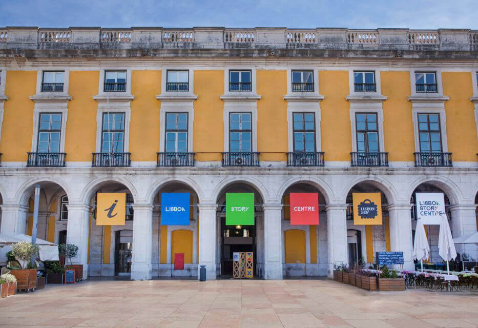 The Lisbon Story Centre, housed in a building with a yellow facade in Praça do Comércio. The entrance features colorful signs for information and exhibitions.