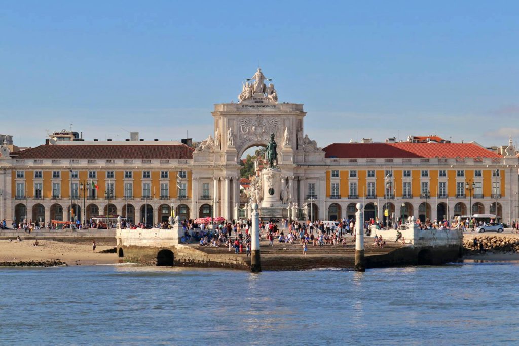 A view of Praça do Comércio from the Tagus River, showcasing the large equestrian statue and the surrounding yellow buildings. The archway of Rua Augusta is visible, with many people gathered around the square.