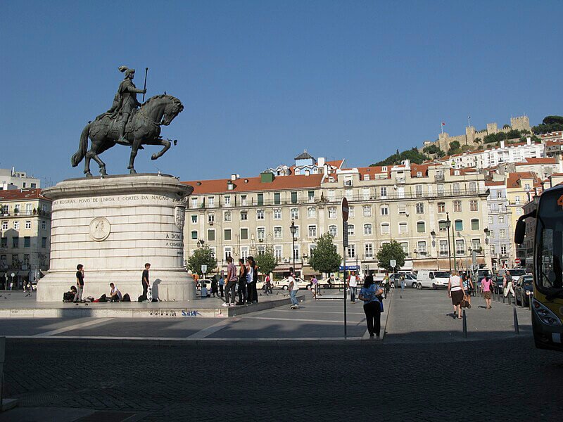 View of Praça da Figueira in Lisbon, featuring the equestrian statue of King João I. The background includes a row of historical buildings and the São Jorge Castle perched on the hill.