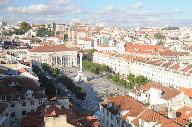 An aerial view of Rossio Square (Praça Dom Pedro IV) in Lisbon. The square features a central monument, surrounded by historical buildings with terracotta roofs, and tree-lined streets.