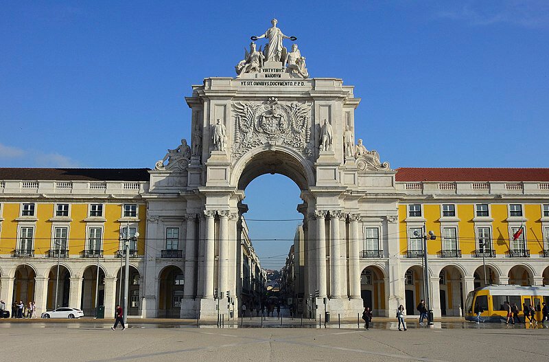 The Rua Augusta Arch, a majestic triumphal arch located at the entrance of Praça do Comércio. The arch is adorned with statues and intricate carvings, with the yellow buildings of the square flanking either side.