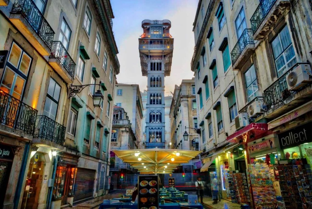 The Elevador de Santa Justa, a neo-Gothic iron elevator in Lisbon. It is framed by narrow streets with historical buildings, and the area below is bustling with shops and cafes.