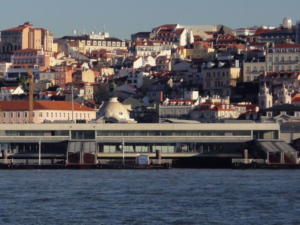 A scenic view of Lisbon's hillside neighborhood, featuring a mix of colorful buildings with terracotta roofs. The foreground showcases modern architecture near the waterfront.