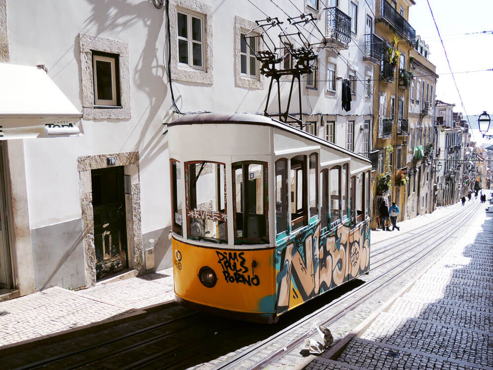 A historic tram covered in graffiti ascends a narrow, cobblestone street lined with traditional buildings in Lisbon.