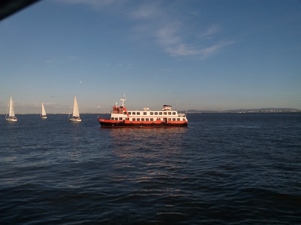 A red and white ferryboat sails on a calm river with several sailboats in the background under a clear blue sky.