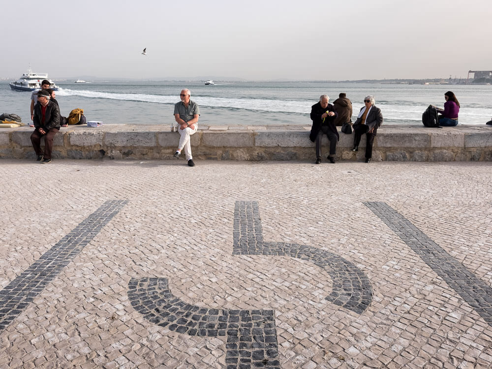 People sitting on a stone ledge by the riverbank, with a mosaic cobblestone walkway in the foreground and boats on the water in the background.