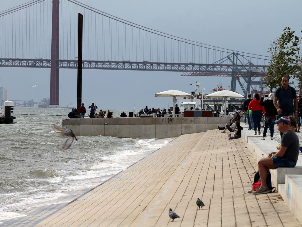 People enjoying a stroll along a waterfront promenade, with the 25 de Abril Bridge visible in the background. The scene includes pigeons, some people sitting, and others walking by the river.