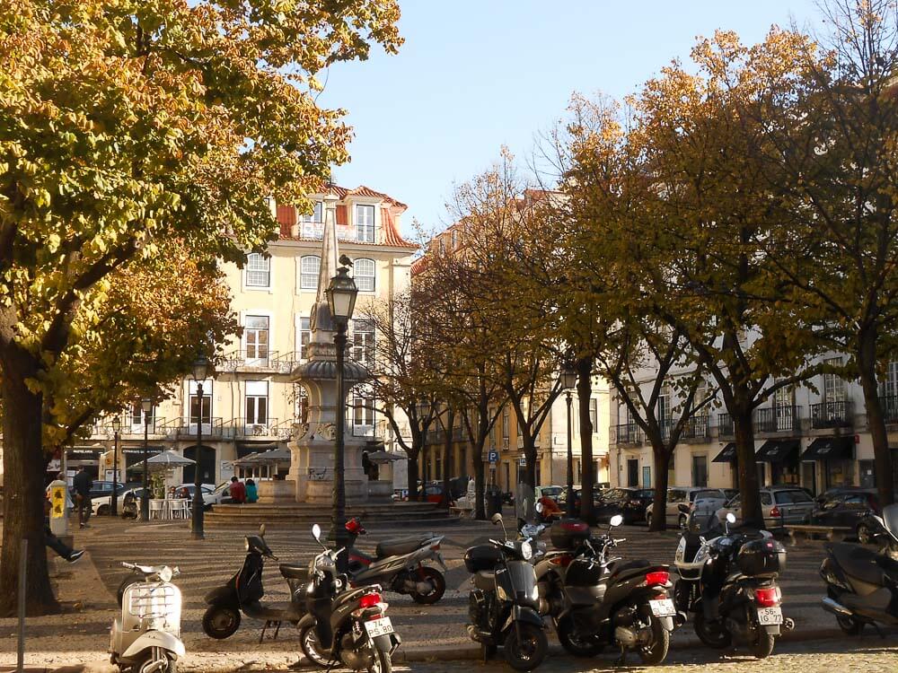 A picturesque square in Lisbon with a central fountain, surrounded by trees and historical buildings. The foreground shows parked scooters and people enjoying the outdoor cafes.