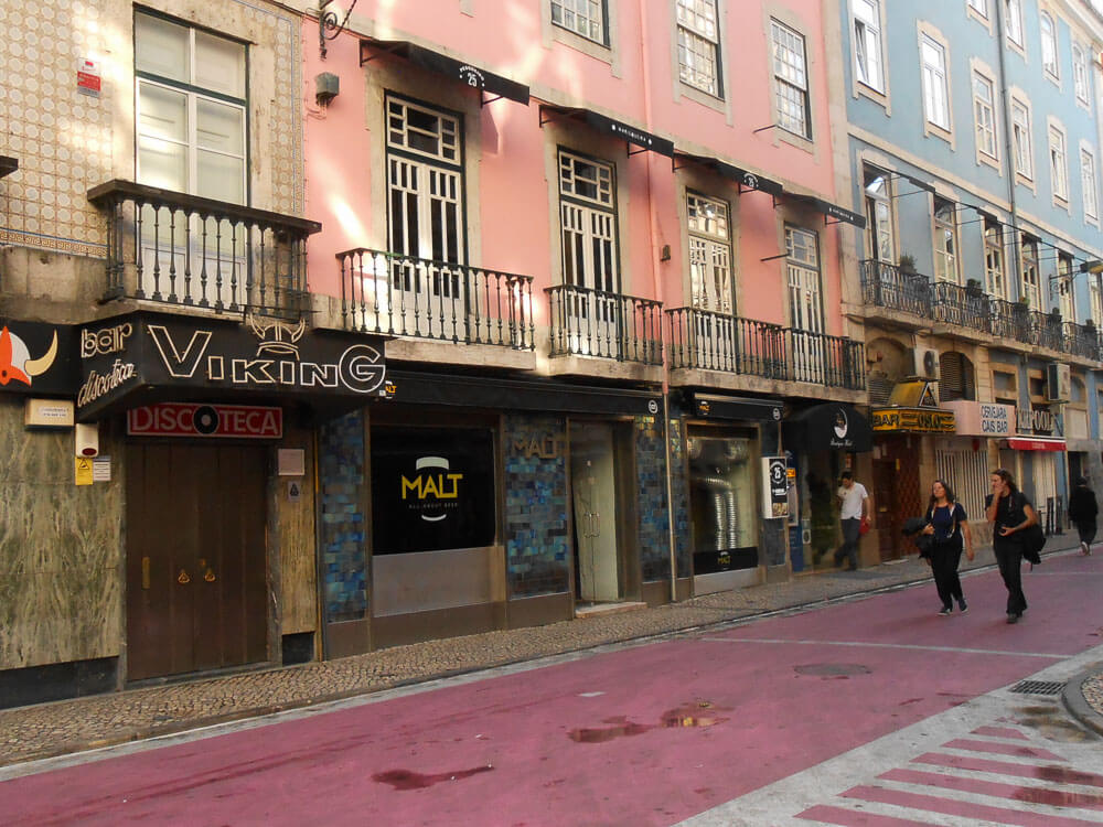 A street in Lisbon lined with colorful buildings featuring bars and shops. The pink-painted street contrasts with the pastel hues of the buildings. Pedestrians are walking along the sidewalk.