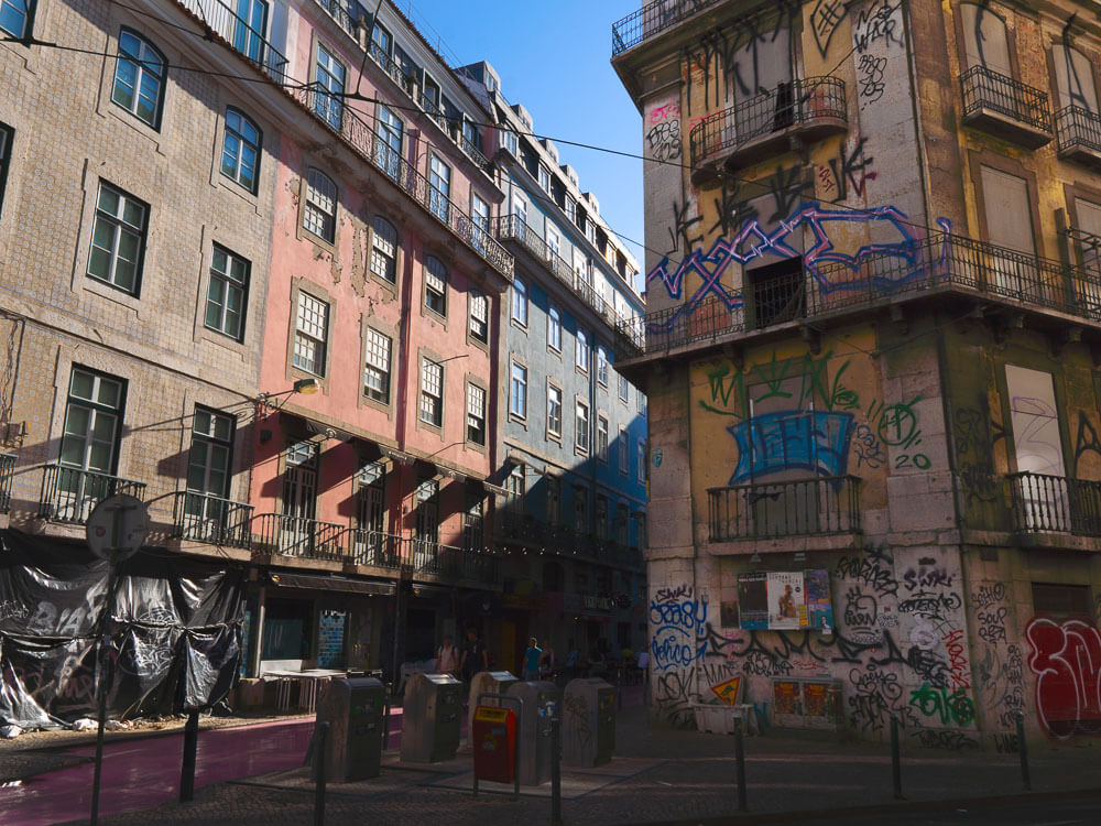 A street corner in Lisbon with buildings covered in graffiti. The architecture features traditional balconies and various colorful street art, adding an urban feel to the scene.