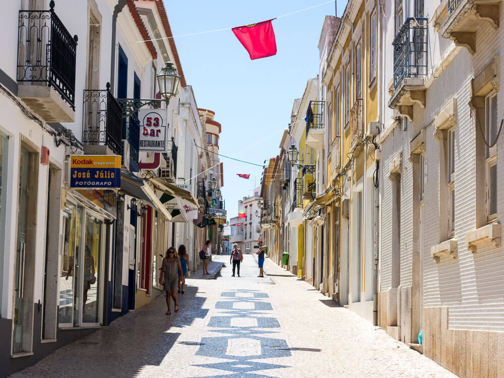 A charming street in Lisbon lined with shops and restaurants, featuring cobblestone pavement with decorative patterns and a few people walking along.