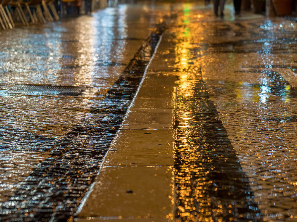 A close-up view of a wet cobblestone street at night, reflecting the lights from nearby buildings and creating a glowing effect.