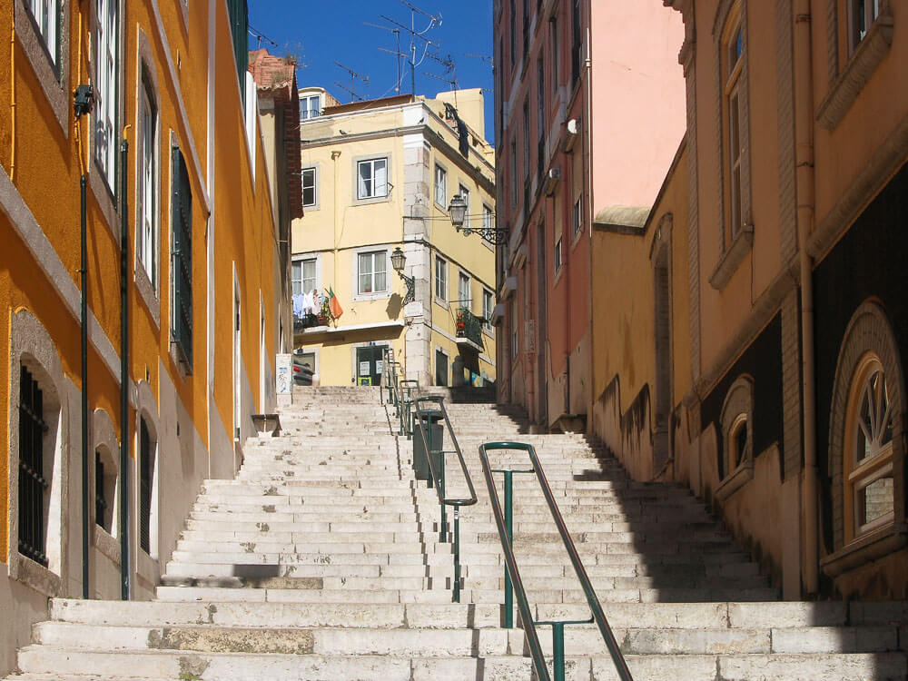 A steep flight of stairs in Lisbon's historical neighborhood, flanked by colorful buildings and leading up to a sunny terrace.