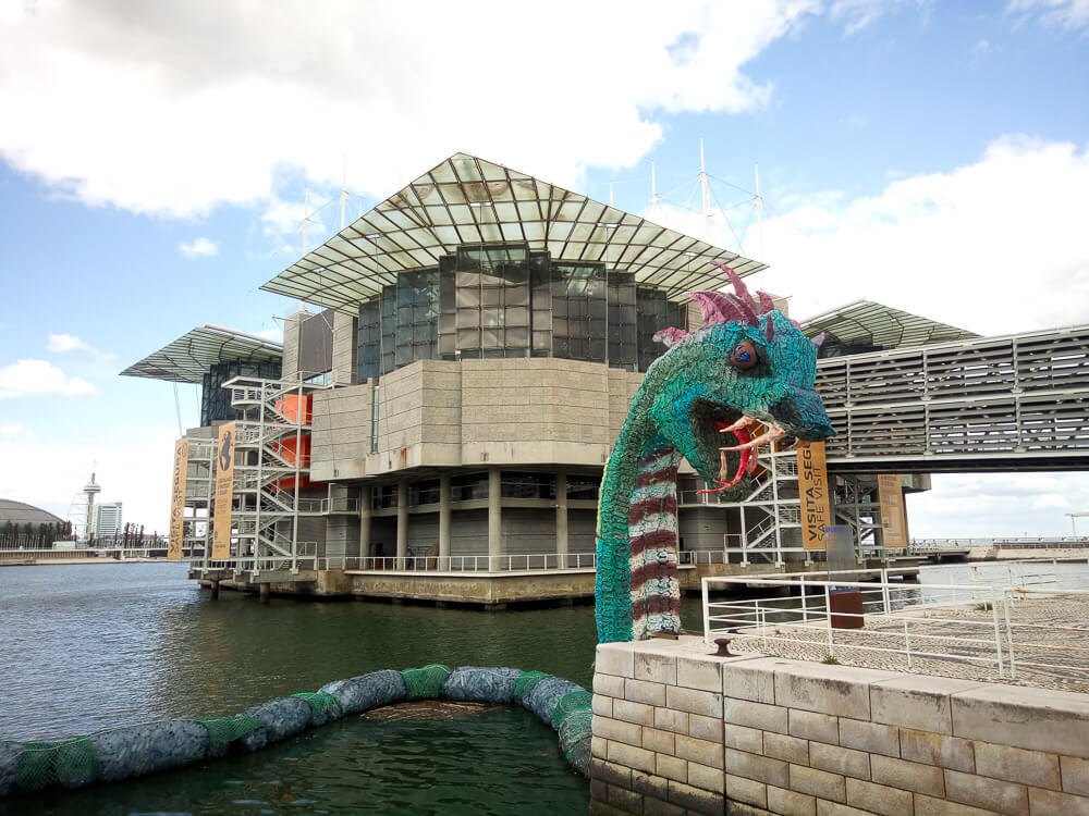 The modern and striking architecture of the Lisbon Oceanarium, seen from the waterfront, with a playful sea creature sculpture in the foreground.