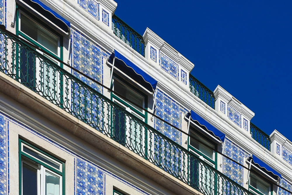 The façade of a Lisbon building adorned with traditional blue and white azulejo tiles, highlighted by green iron balconies and blue awnings.