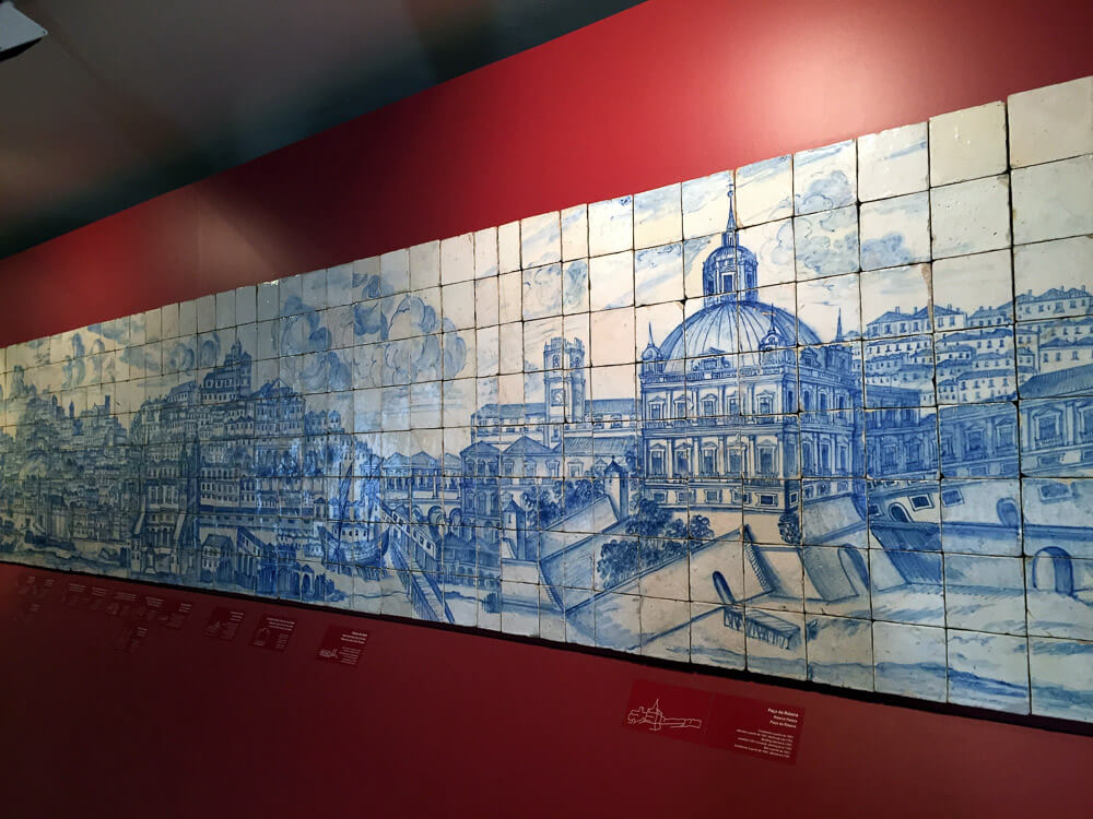 A large azulejo mural depicting a historical cityscape of Lisbon, displayed on a red wall, showcasing intricate details and panoramic views.