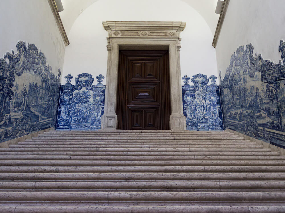 A grand staircase adorned with intricate blue azulejo tile murals, leading to an imposing wooden door in a historic Lisbon building.