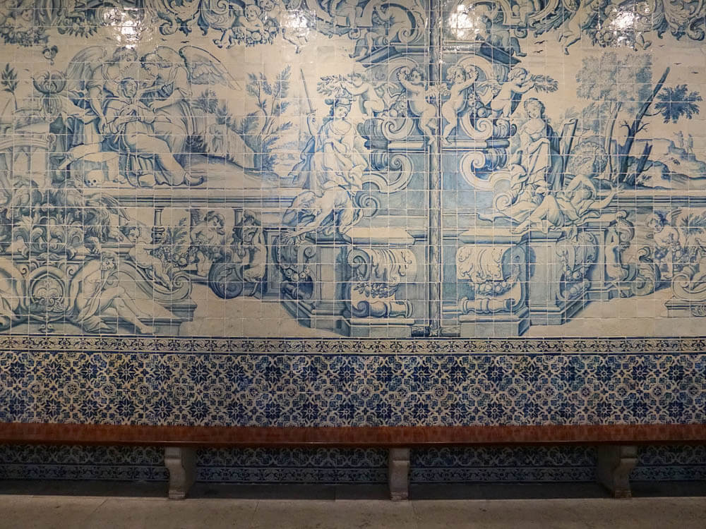 A detailed azulejo mural in Lisbon, depicting classical scenes with cherubs and elaborate architectural elements in shades of blue and white.