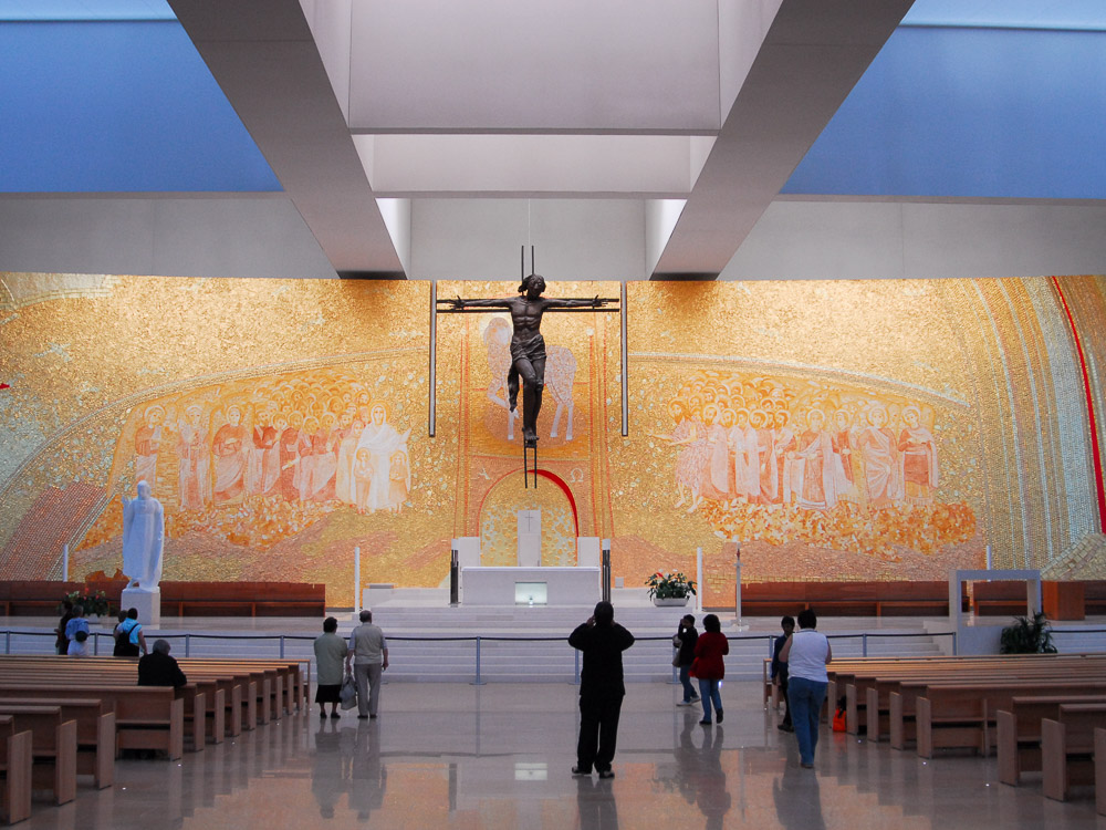 The interior of a modern church with a large golden mosaic depicting religious figures behind the altar, and a suspended crucifix in the center. Visitors are seen admiring the artwork.