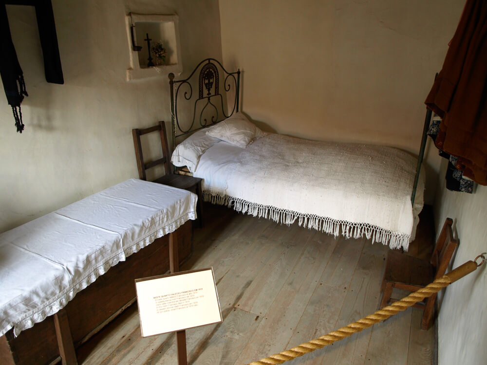 A simple bedroom with rustic furnishings, including a bed with a wrought-iron headboard, a wooden bench, and a small chair. The room is part of a historical exhibit.