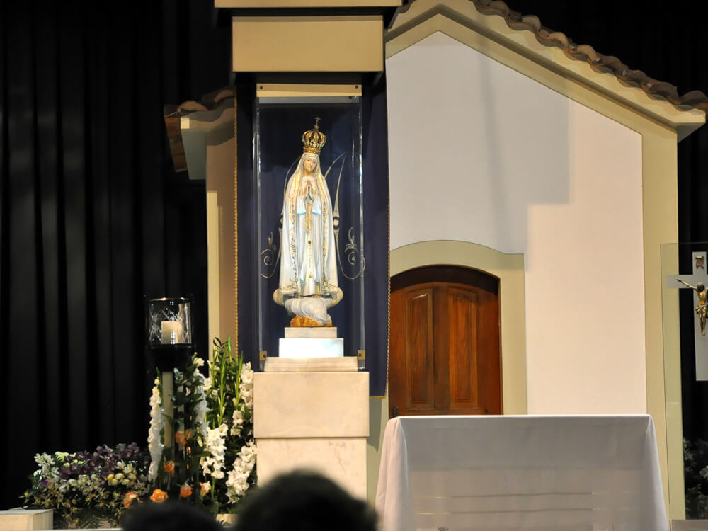 A statue of Our Lady of Fátima inside a chapel, adorned with a crown and surrounded by floral decorations, placed on an altar.