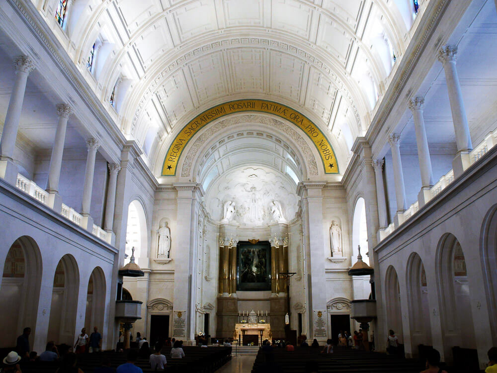 The interior of the Basilica of Our Lady of the Rosary in Fátima, featuring a white vaulted ceiling, large archways, and religious statues.