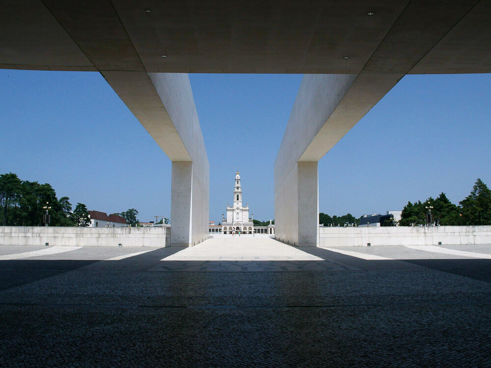 A view through a modern architectural structure framing the distant Sanctuary of Our Lady of Fátima, with its iconic bell tower.