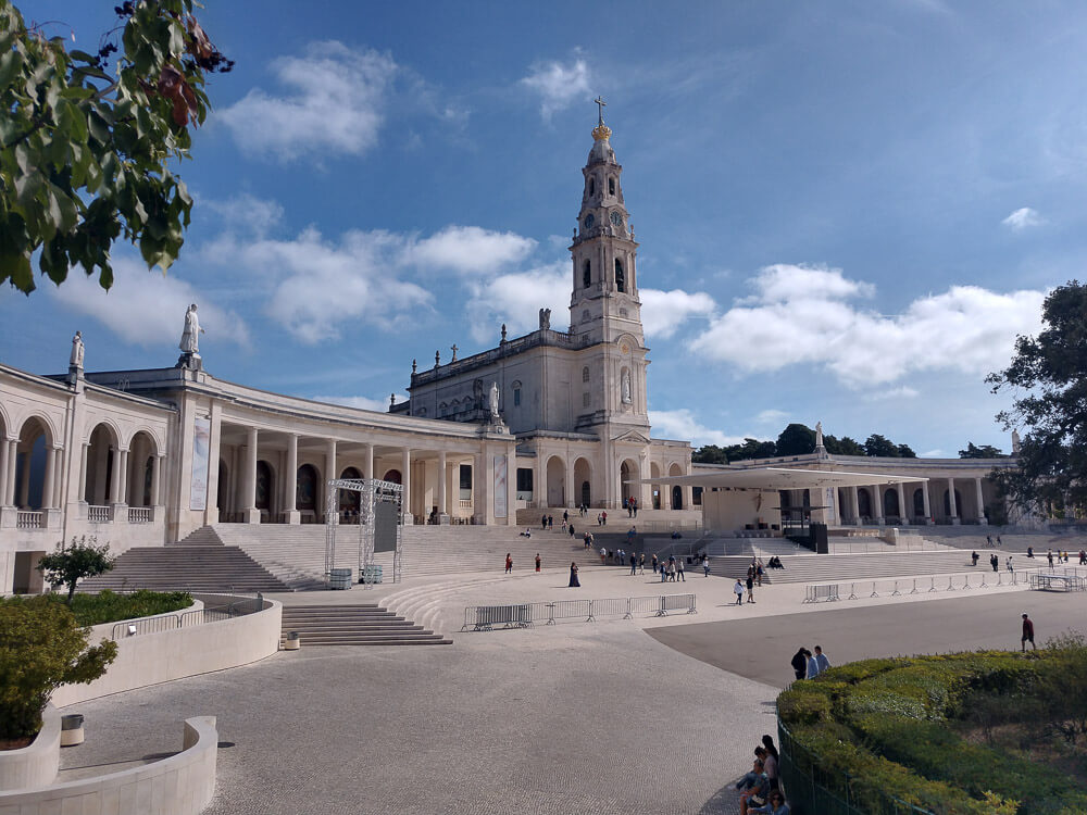 An expansive view of the Sanctuary of Our Lady of Fátima, including the central basilica and the surrounding colonnades, with visitors walking around the large plaza.