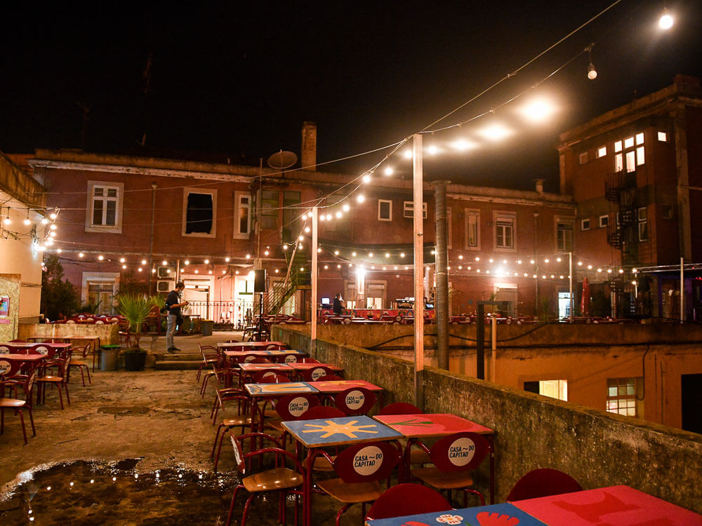 Outdoor seating area of a bar in Marvila, Lisbon, decorated with colorful tables and illuminated by string lights at night.