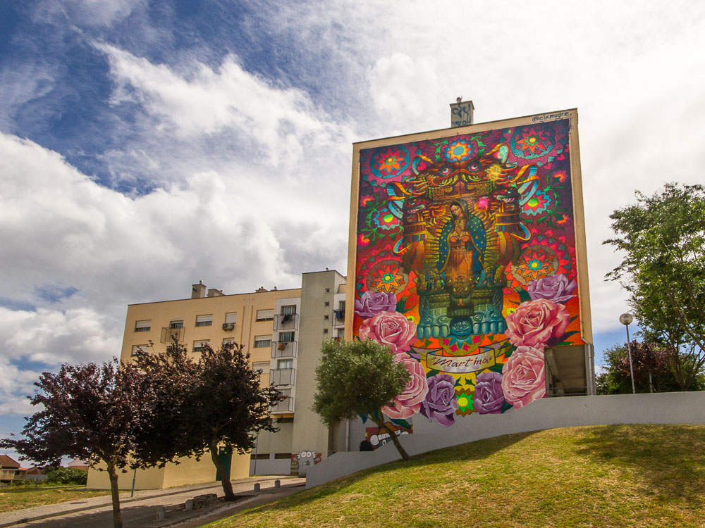 Large, colorful mural on the side of a building in Marvila, Lisbon, depicting intricate designs and a central figure, with a clear sky above.