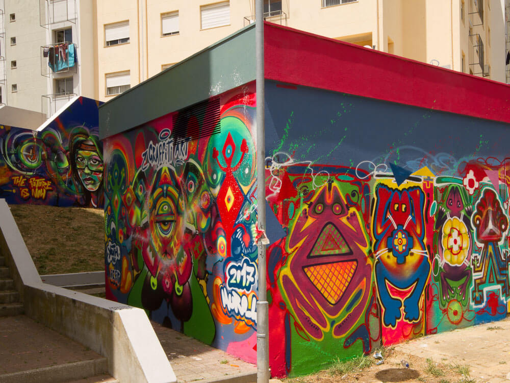 A colorful mural featuring abstract and whimsical designs on a wall in Marvila, Lisbon, with buildings in the background.