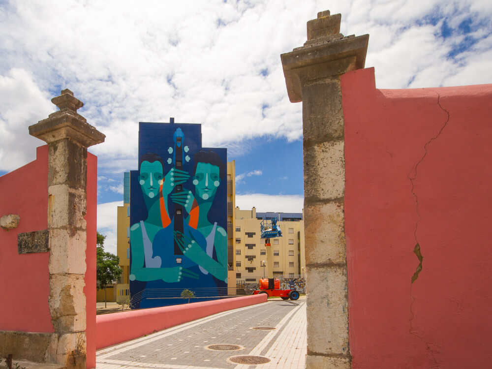 View through a pink archway in Marvila, Lisbon, framing a tall mural of two blue-toned figures on a building wall.