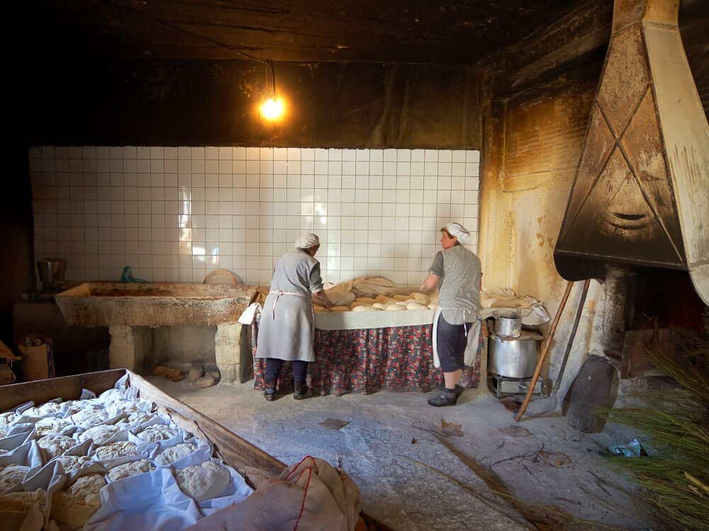 Two people work in a rustic kitchen kneading dough on a table. The room has a stone sink, tiled wall, and hearth. Various baking supplies and dough awaiting preparation are visible.