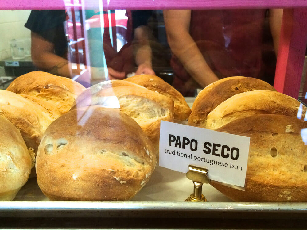 Bread arranged on a tray. There is a sign saying "papo seco (traditional Portuguese bun)" and two people in the background.