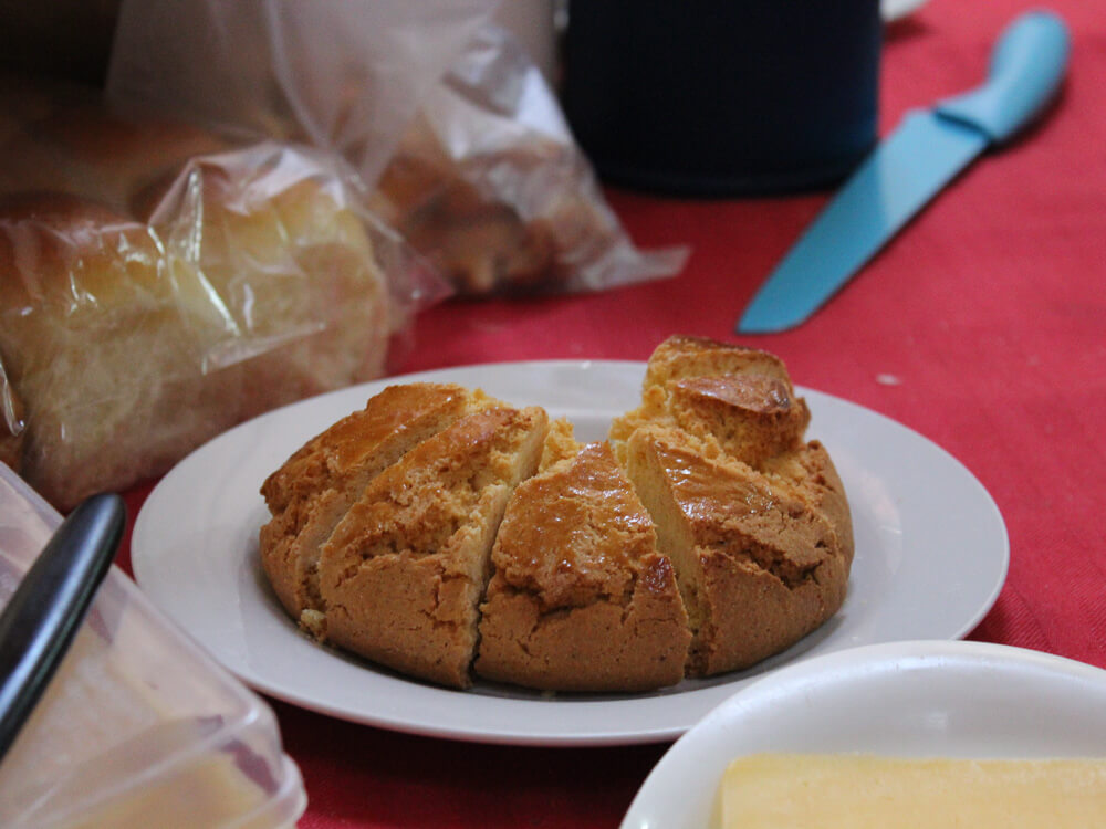 A deep white plate on top of a red tablecloth. On it, there is a corn bread cut into pieces. In the background, a knife and a transparent package with bread.