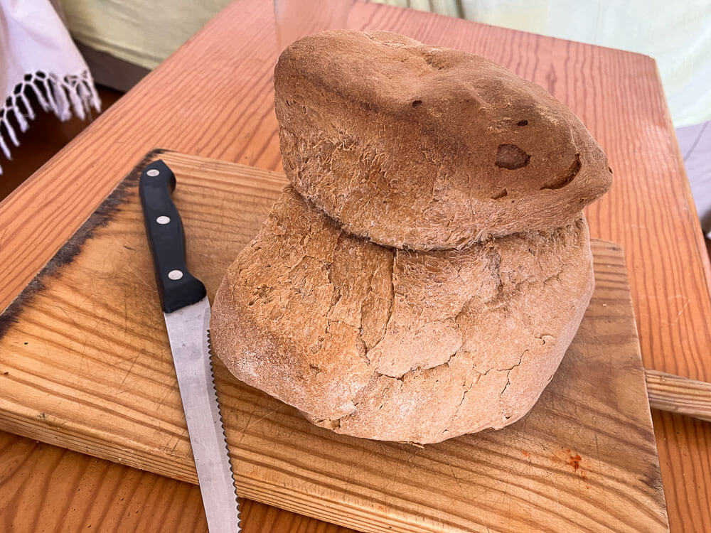 A wooden board on a wooden table. On it, there is a bread knife and an Alentejo bread.