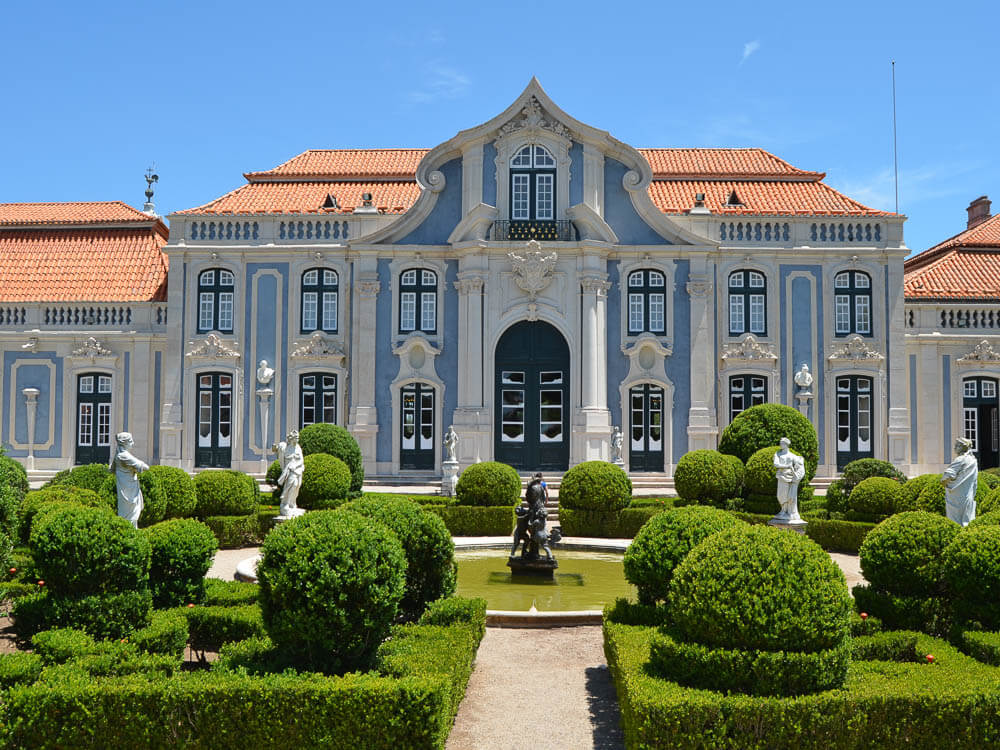 The grand façade of Queluz Palace, featuring elegant blue and white architecture, with beautifully manicured gardens and classical statues in the foreground.