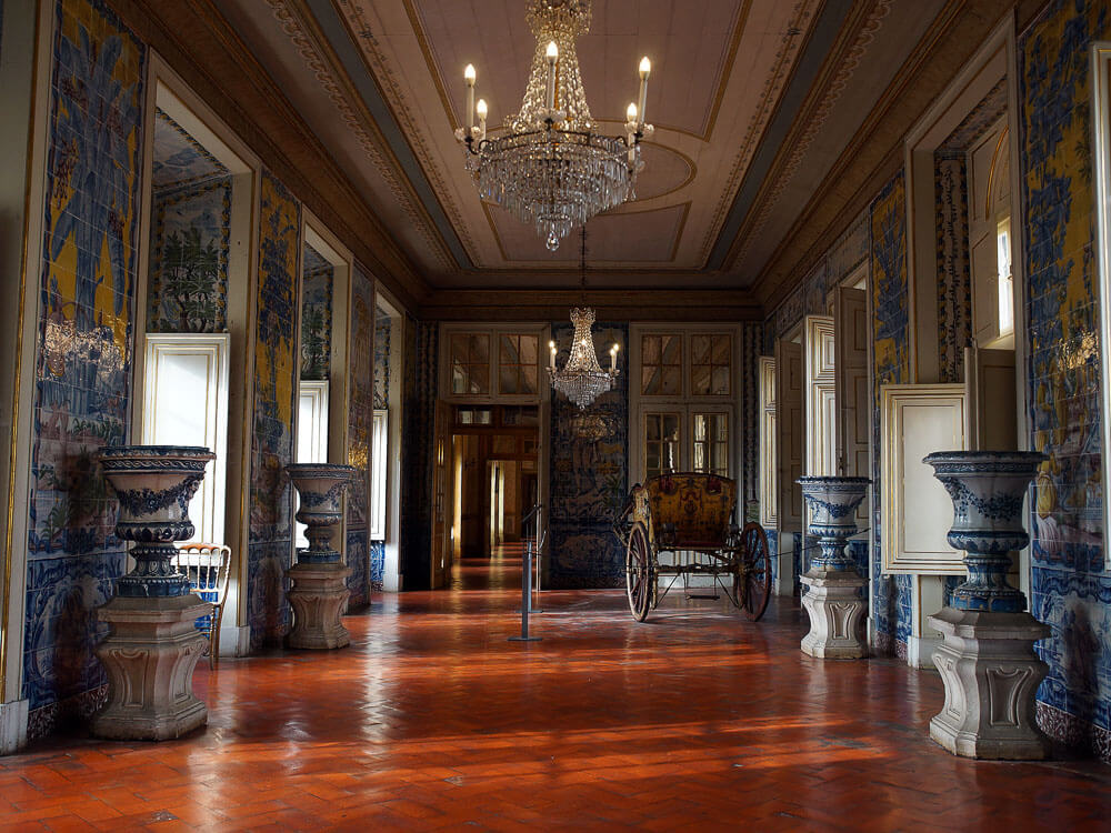 A luxurious interior hallway of Queluz Palace, adorned with intricate blue and white tile murals, opulent chandeliers, and a historic carriage.