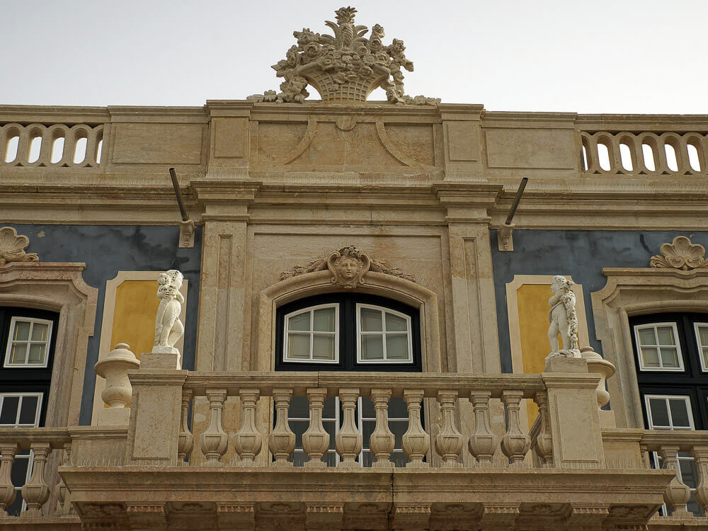 A close-up of the ornate balcony and detailed stonework on the façade of Queluz Palace, showcasing classical architectural elements and sculptures.