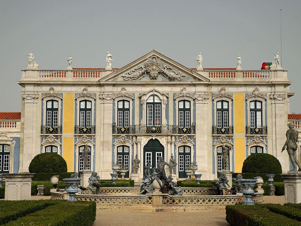 The majestic front view of Queluz Palace, highlighting its grand entrance, intricate details, and symmetrical design, framed by a lush garden with statues and fountains.