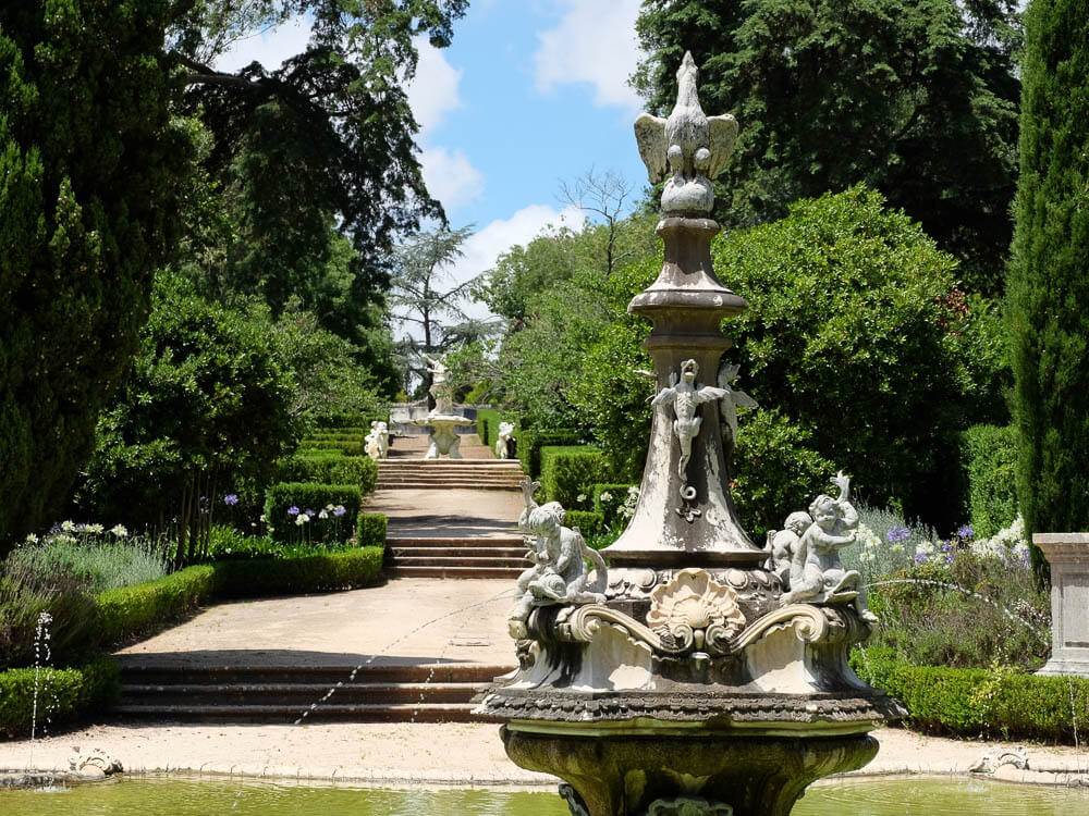 A charming garden fountain at Queluz Palace, surrounded by lush greenery and leading up to a staircase lined with classical statues.