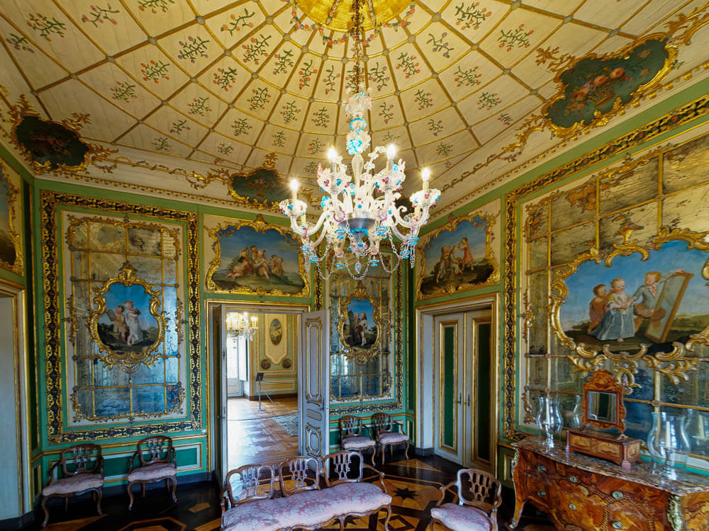 A richly decorated room inside Queluz Palace, featuring an ornate ceiling, lavish chandeliers, and walls adorned with elaborate paintings and gold accents.