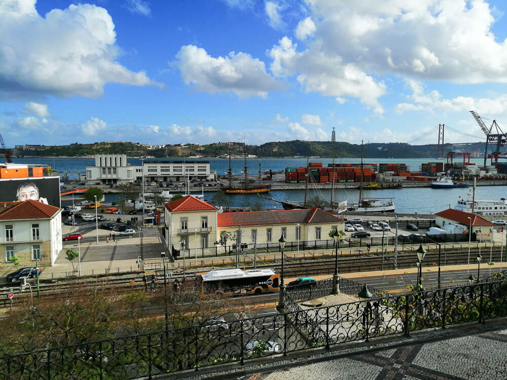 A view of Lisbon's port area, featuring the 25 de Abril Bridge, numerous shipping containers, and cranes along the waterfront.