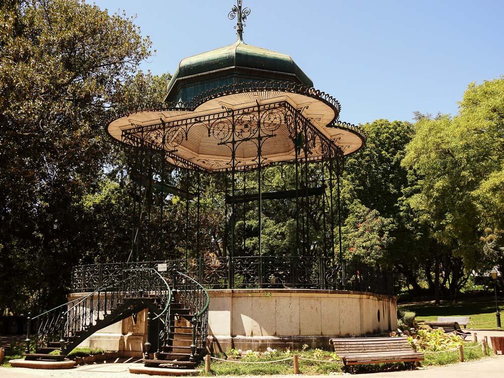 A gazebo in a Lisbon park, featuring ornate wrought-iron details and surrounded by lush greenery and benches.