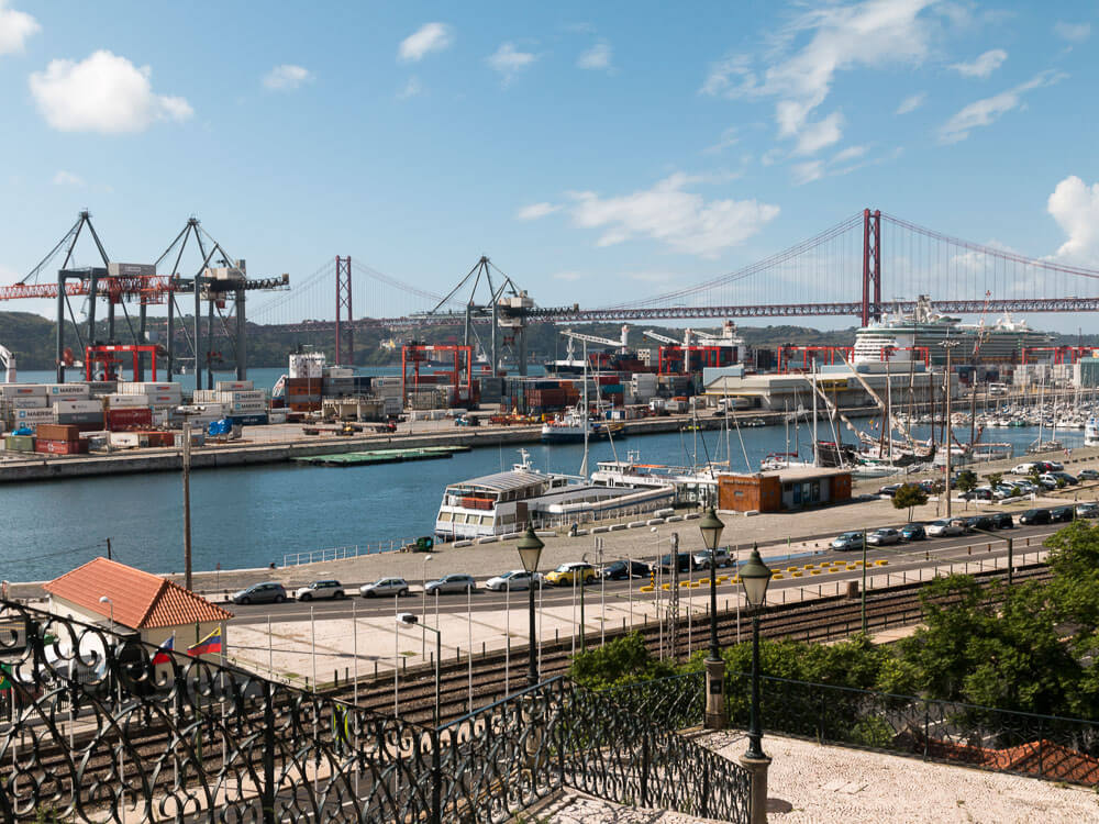 A picturesque view of Lisbon's waterfront area, with boats docked at the marina, the 25 de Abril Bridge in the background.