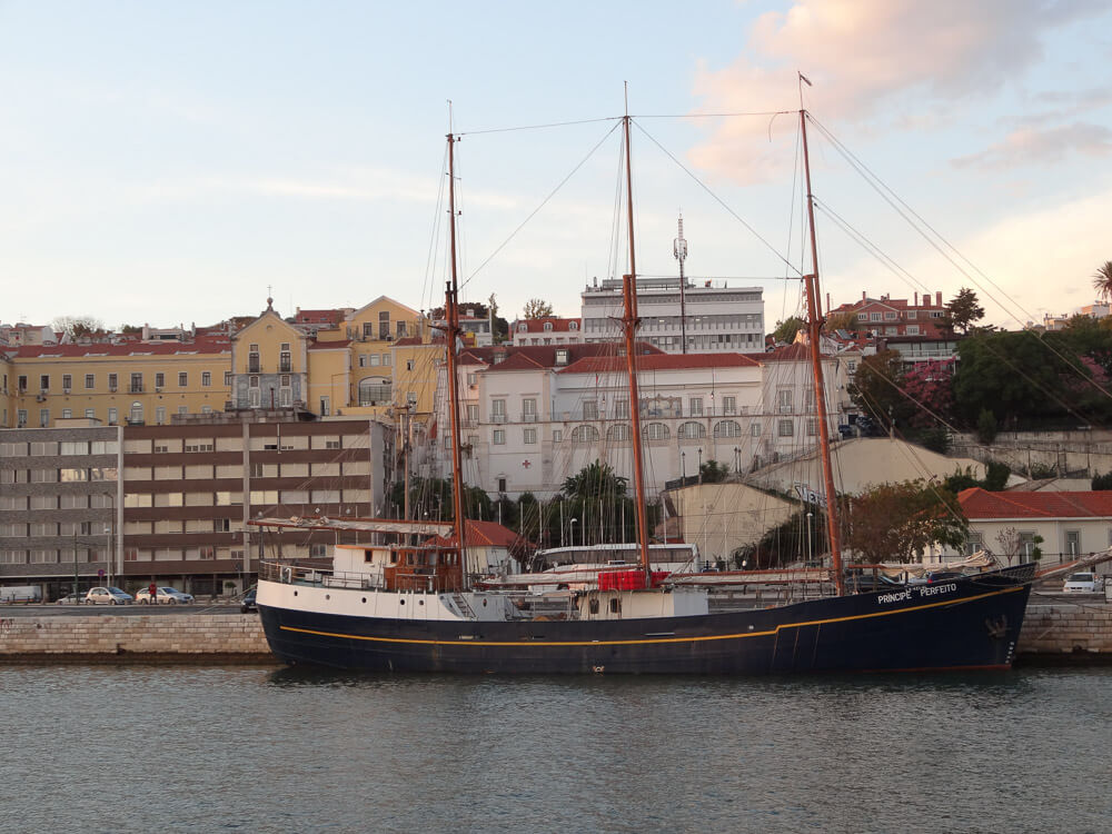 A large sailboat docked in Lisbon's harbor, with a backdrop of historic buildings and the skyline of the city.