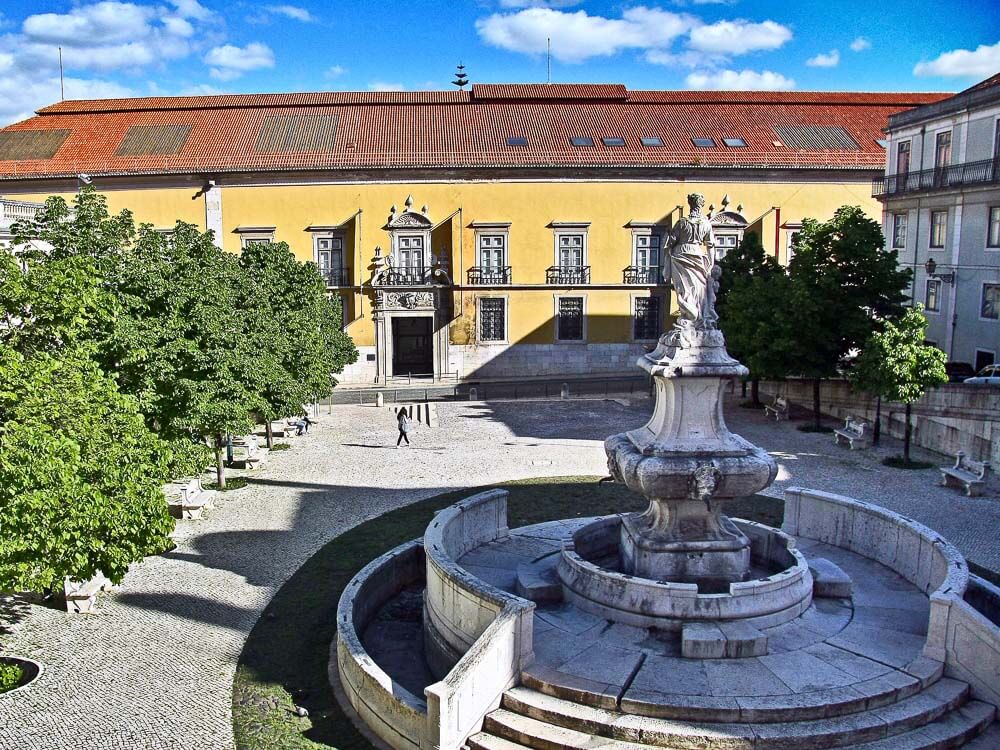 The courtyard of a historical building in Lisbon, featuring a central fountain, manicured trees, and a yellow facade with arched windows.